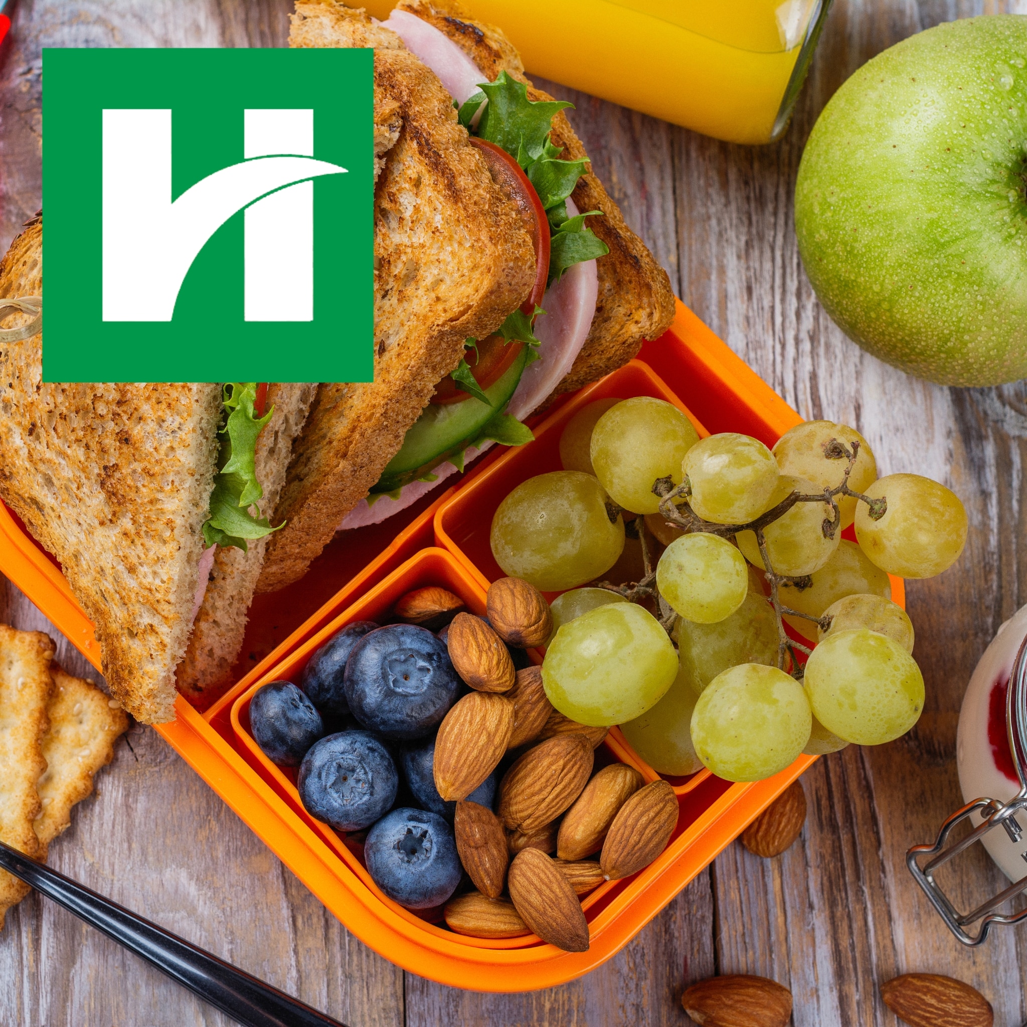 Orange lunchbox filled with a sandwich, grapes, blueberries and almonds, with a green apple and orange juice nearby and the Hendricks Regional Health logo in the upper left.