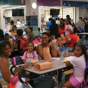 Families sit together at tables enjoying food and bottled water during a busy YMCA Back to School Fest, with children wearing backpacks and smiling for the camera.