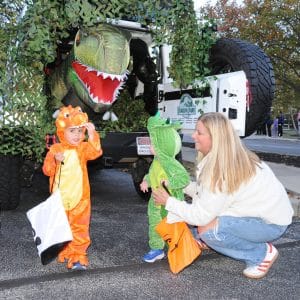 Children in dinosaur costumes trick-or-treat beside a Jurassic Park-themed vehicle display during a YMCA Fall Fest family event.