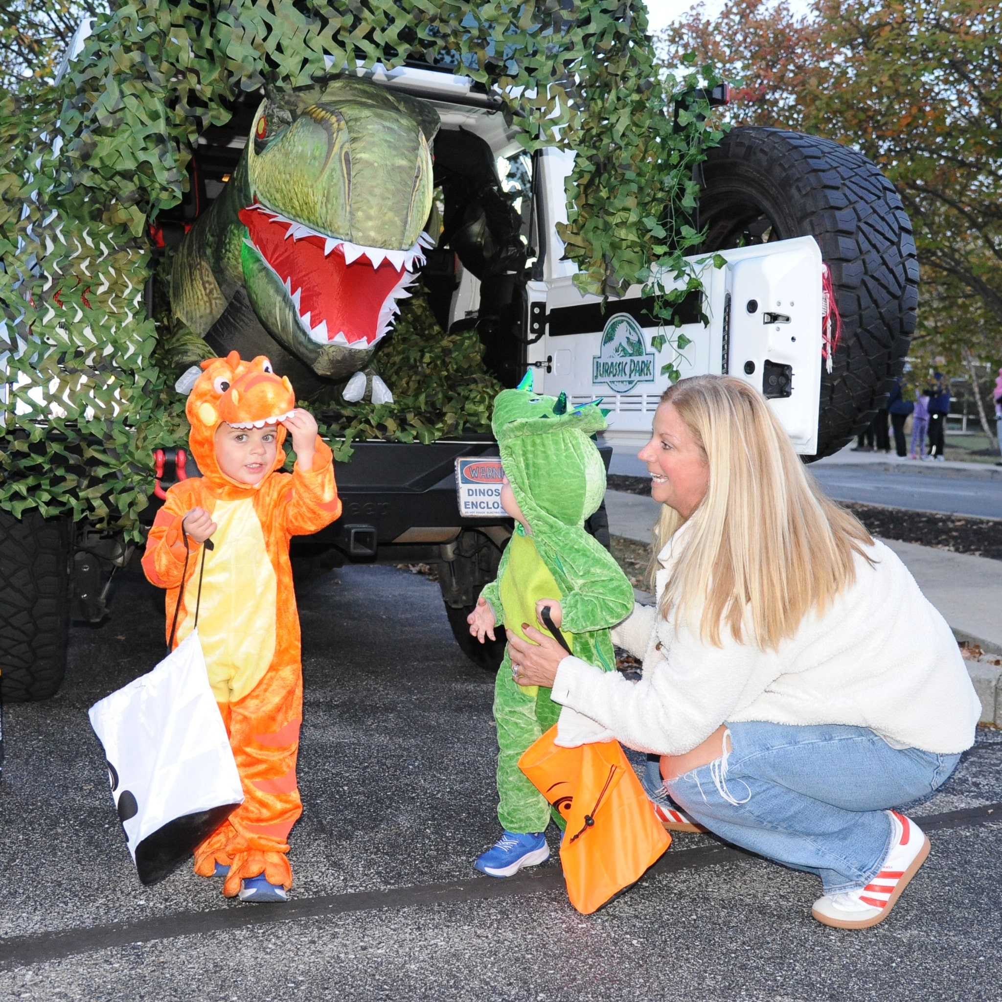 Children in dinosaur costumes trick-or-treat beside a Jurassic Park-themed vehicle display during a YMCA Fall Fest family event.