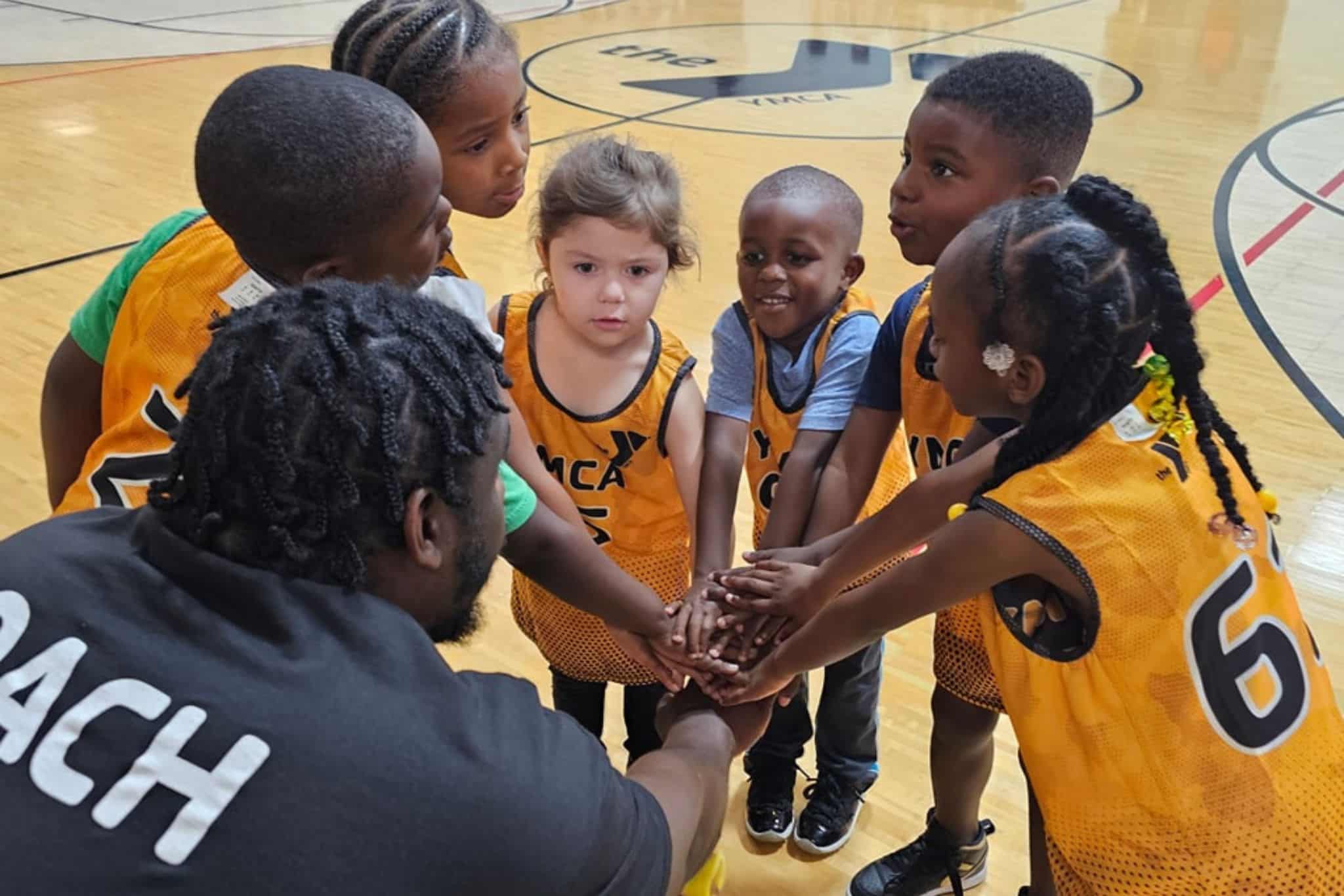 preschool children in yellow sport uniforms standing in a circle with a coach and placing their hands together