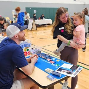 Volunteer at a YMCA Healthy Kids Day event smiles and chats with a young child being held by a young woman at an information table with pens, stickers and water bottles, while families explore activities in a gymnasium in the background.