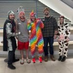 Five YMCA staff members pose indoors in Halloween costumes for Trick-or-Treating at the Y, including a knight, shark, rainbow unicorn and cow.