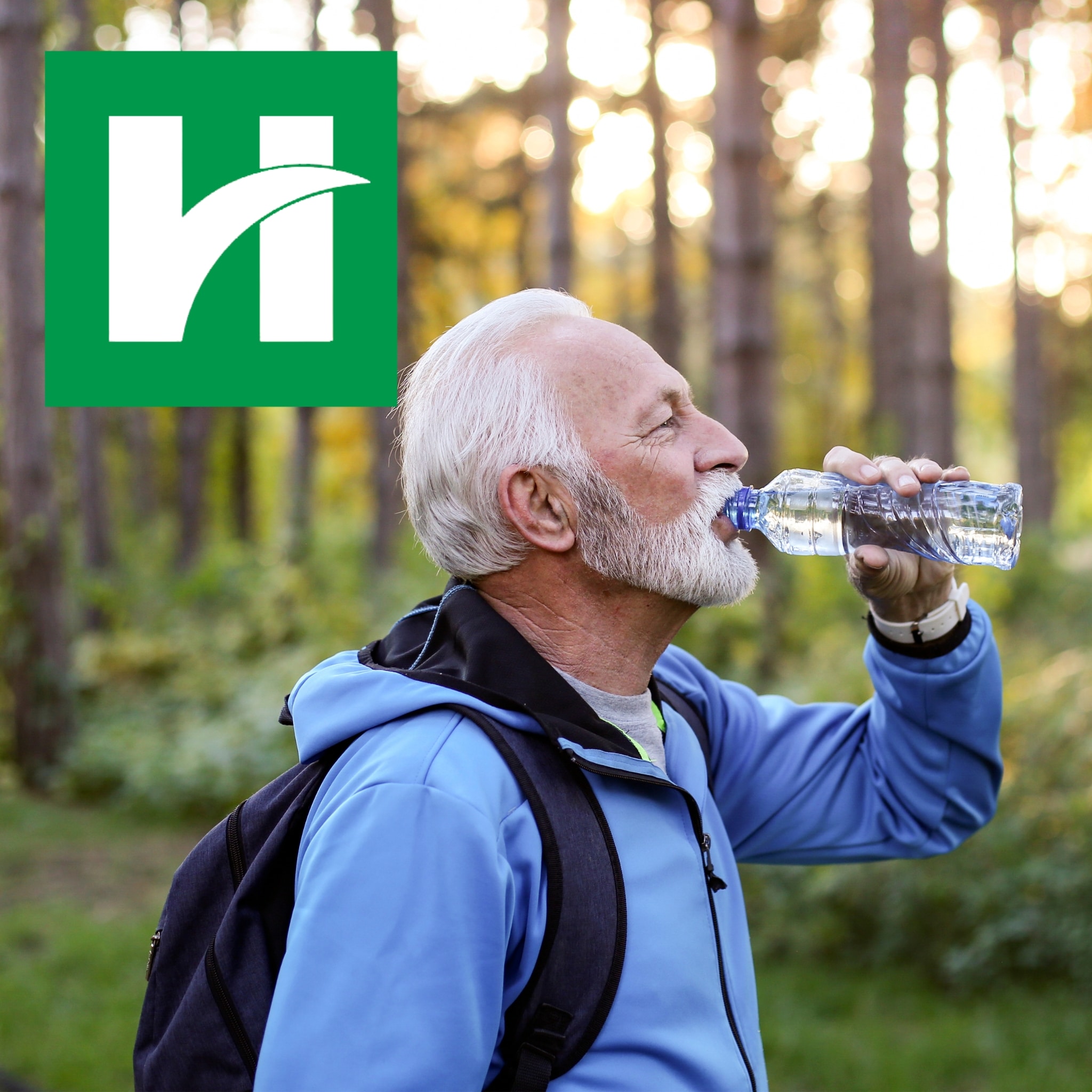 Older man in a blue jacket drinks water from a bottle while hiking in a wooded area, with the Hendricks Regional Health logo in the upper left corner.